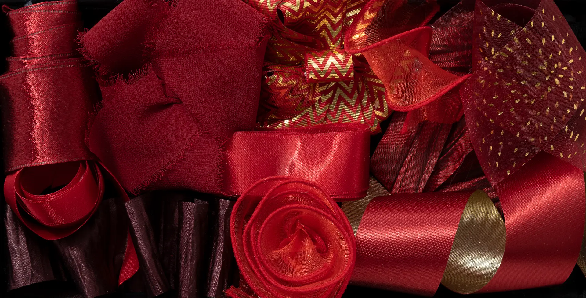 A high-detail studio photograph of various red and gold ribbons in different textures, including satin, grosgrain, and frayed silk. The composition features a mix of solid reds, metallic gold zig-zag patterns, and shimmering gold-flecked fabrics arranged in a dense, tactile from overhead