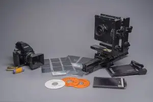 A professional studio still life featuring vintage and large-format photography equipment on a grey background. The scene includes a Zenza Bronica medium-format camera, a Horseman large-format rail camera with bellows, rolls of 120 film, a Polaroid film holder, transparency contact sheets, and three labeled DVDs dating from 2002 and 2003.