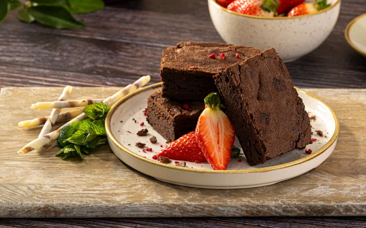 A stack of thick, fudgy chocolate brownies garnished with fresh strawberry slices, freeze-dried berry crumbles, rolled wafer curls, and fresh mint leaves on a round plate.