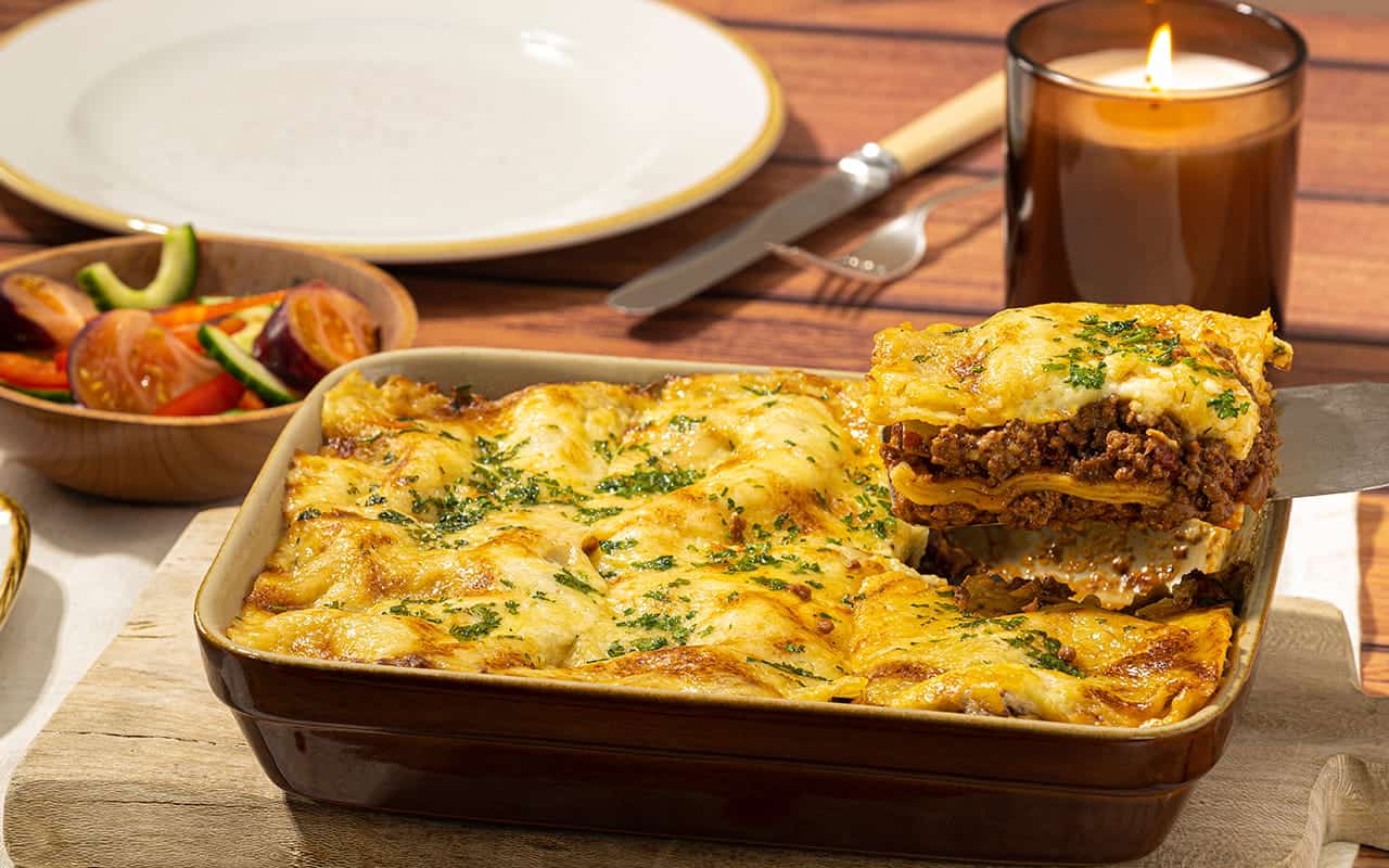 A spatula lifting a slice of beef lasagna from a ceramic baking dish, revealing layers of minced beef, pasta sheets, and melted cheese, served alongside a side salad on a rustic wooden table
