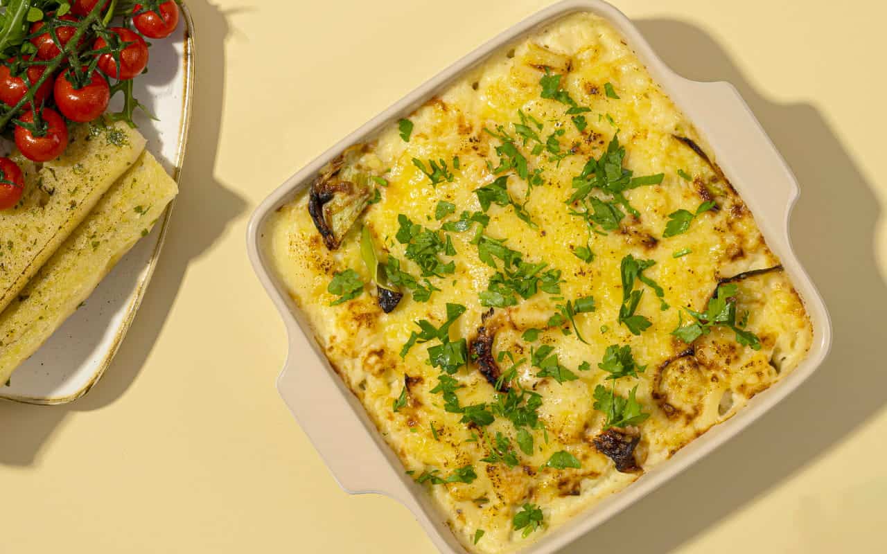 A square baking dish filled with golden baked macaroni cheese topped with chopped fresh parsley, served next to garlic bread and roasted vine tomatoes on a light yellow background.