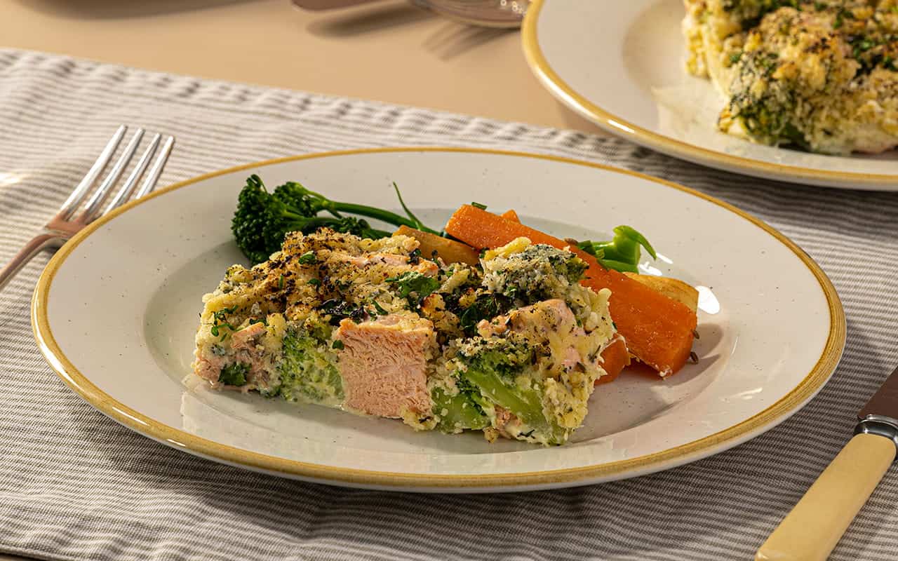 A slice of baked salmon and broccoli casserole topped with a golden herb crust, served with roasted carrots and tenderstem broccoli on a ceramic plate.